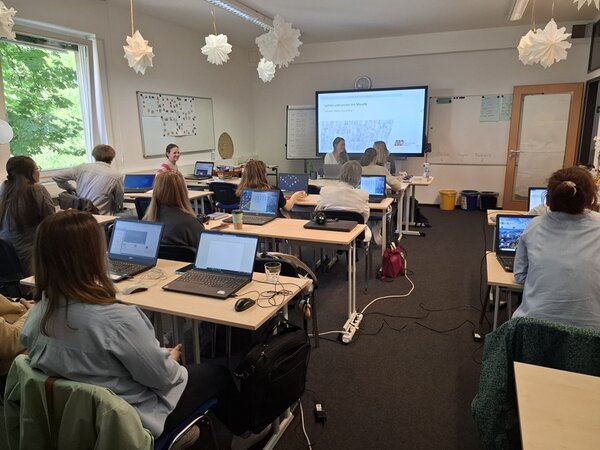 Mitarbeitende der Euro-Schulen sitzen zur Weiterbildung in einem großen Unterrichtsraum an Tischen, über ihnen hängen weiße Papiersterne, grauer Teppichboden ist ausgelegt. Vorn wird eine Präsentation am großen Bildschirm gezeigt.