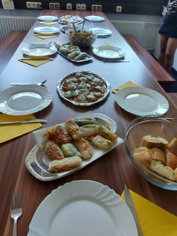 Ein Esstisch ist für mehrere Personen gedeckt. Auf dem Tisch stehen verschiedene Speisen, darunter gefüllte Röllchen, ein Salat und Brot. Gelbe Servietten und weiße Teller sind bereitgestellt.