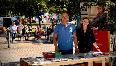 Ein Mann und eine Frau stehen an einem Infostand in einem Hof. Auf dem Tisch liegen Broschüren, dahinter steht ein Roll-up der Euro-Schulen. Im Hintergrund sieht man Menschen an Tischen sitzen, internationale Fahnen sind an einer Schnur aufgehängt. Es ist sonnig.
