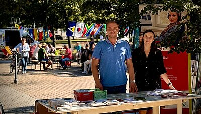 Ein Mann und eine Frau stehen an einem Infostand in einem Hof. Auf dem Tisch liegen Broschüren, dahinter steht ein Roll-up der Euro-Schulen. Im Hintergrund sieht man Menschen an Tischen sitzen, internationale Fahnen sind an einer Schnur aufgehängt. Es ist sonnig.