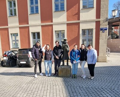 Mehrere Teilnehmende der Euro-Schulen Bamberg stehen nebeneinander auf einem gepflasterten Platz vor einem historischen Gebäude mit rot-weißer Fassade. In der Mitte der Gruppe befindet sich eine bronzefarbene Statue auf einem Sockel. Die Personen tragen überwiegend dunkle Jacken und Hosen, eine Person trägt eine helle Jacke. Im Hintergrund sind ein kleines schwarzes Auto und ein weiteres Fahrzeug zu sehen. Rechts an der Wand hängt ein blaues Straßenschild.