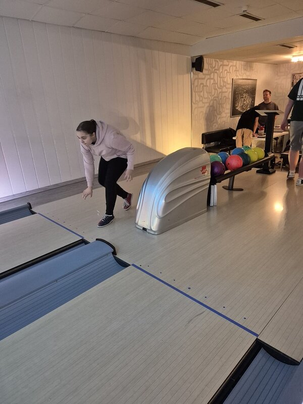 Eine Teilnehmerin im hellen Hoodie geht beim Bowling in die Wurfbewegung und beugt sich nach vorn; rechts daneben stehen zahlreiche farbige Kugeln auf dem Kugelständer.