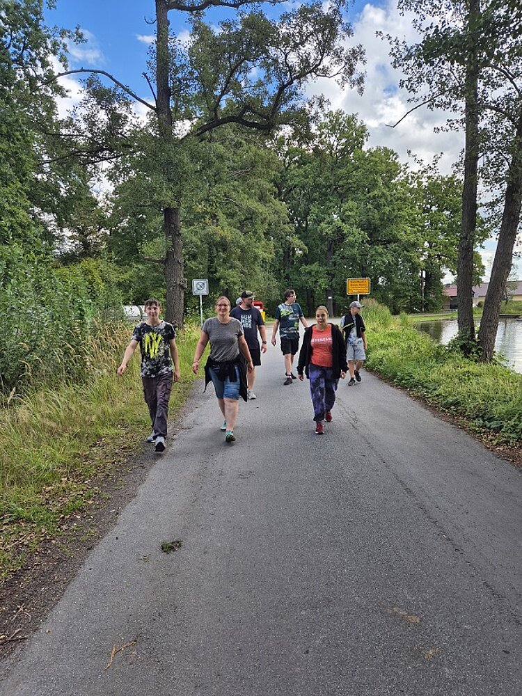 Gruppe von Jugendlichen läuft auf einer Landstraße durch eine grüne Waldlandschaft, im Hintergrund ein Ortsschild und ein Fluss.