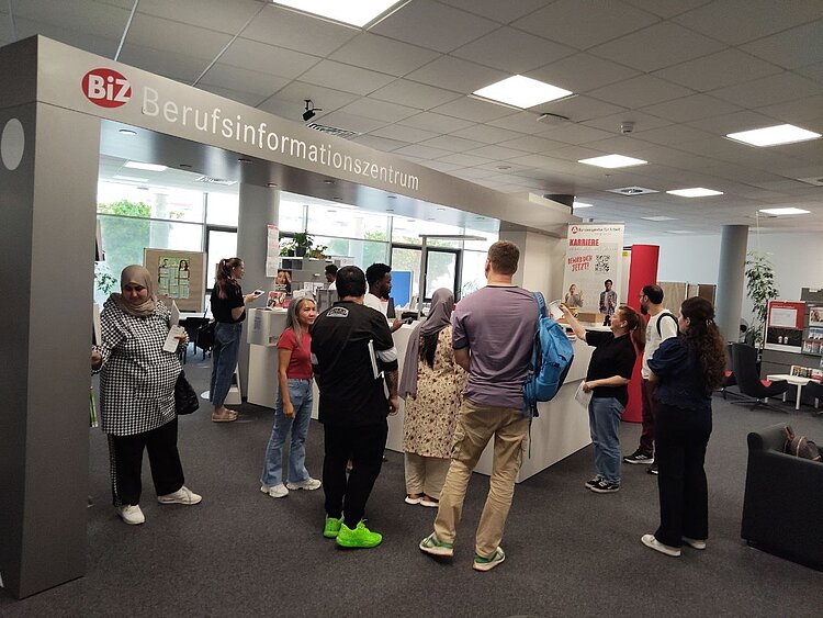 Gruppe IK-359 zu Besuch im BIZ Eine Gruppe von Teilnehmenden des Integrationskurses steht im Berufsinformationszentrum und erhält Informationen von einer Beraterin, die ein Dokument hochhält..