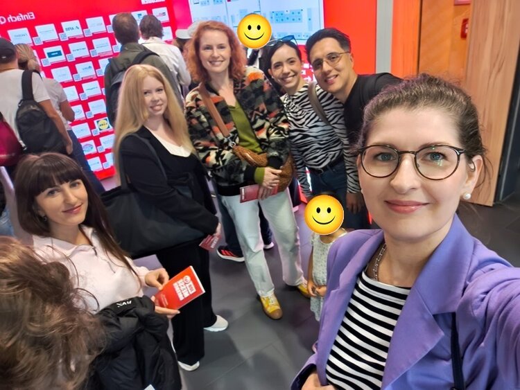 Eine Gruppe von Teilnehmenden steht in einem Innenraum vor einer rot-weißen Wand mit zahlreichen rechteckigen Feldern. Im Vordergrund hält eine Person mit Brille und lila Blazer die Kamera für ein Selfie. Die Personen tragen unterschiedliche, teils gemusterte Oberteile und lächeln. Zwei Gesichter sind durch gelbe Smileys verdeckt. Im Hintergrund sind weitere Personen zu sehen, die sich von der Kamera abwenden.