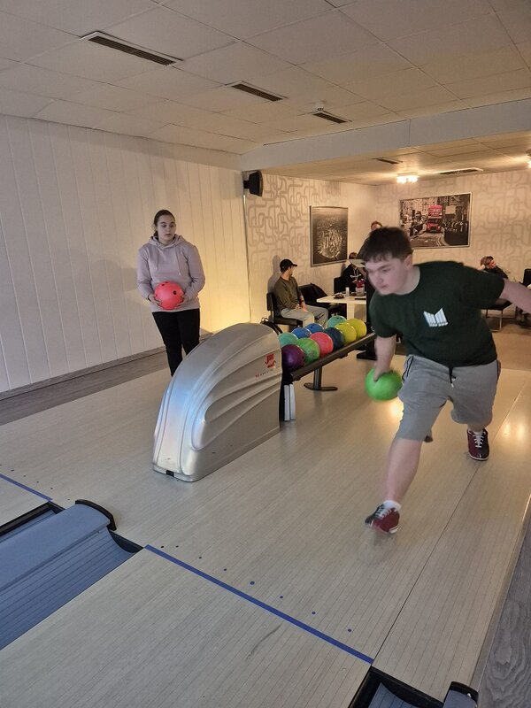 In einer Bowlingbahn rollt ein junger Mann eine grüne Kugel an; hinter ihm wartet eine Teilnehmerin mit roter Kugel, daneben stehen viele bunte Bowlingkugeln auf dem Rücklauf.