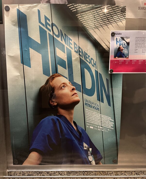 Das Plakat zum Film "Heldin". Zu sehen ist eine Pflegerin in blauer Arbeitskleidung, die erschöpft an einer Aufzugwand lehnt und mit leerem Blick nach oben sieht. Über ihr in baluer Schrift: Leonie Benesch – Heldin