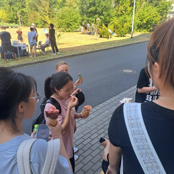 Mehrere Schülerinnen haben Waffeln mit verschiedenfarbigen Eiskugeln in ihrer Hand. Eine junge Frau hält in der anderen Hand ein Smartphone, auf dem das Foto einer Eiswaffel erkennbar ist. Im Hintergrund sieht man zwei Stationen des Sportfestes, wo weitere Personen stehen.