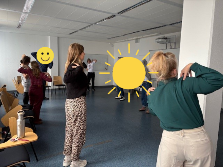 Rück’n’Roll – Bring Bewegung in dein Leben! Die Mitarbeitenden stehen in einem großen Raum und führen, im Rahmen des Gesundheitstages der AOK, Übungen durch. Sie tippen sich mit den Händen stehend auf die Schultern.