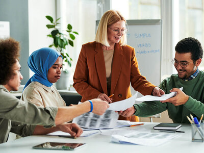 Vier Personen sitzen um einen Tisch in einem Besprechungsraum oder Büro. Eine Frau in einem braunen Blazer steht und gibt Anweisungen oder Erklärungen, während sie auf ein Blatt Papier zeigt. Die anderen drei Personen, darunter eine Frau mit einem blauen Hijab und ein Mann mit einer Brille, hören aufmerksam zu und scheinen Notizen zu machen oder Dokumente zu überprüfen. Auf dem Tisch liegen verschiedene Papiere, ein Tablet und Schreibutensilien. Im Hintergrund ist ein Whiteboard mit Notizen zu sehen, und der Raum ist gut beleuchtet.