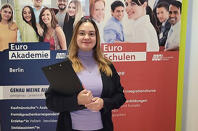 Dozentin Liliia Redko posiert lächelnd für ein Foto. Sie hält ein schwarzes Klemmbrett in der Hand, trägt ein veilchenfarbenen Rollkragenpulli, einen schwarzen Cardigan darüber, eine dünne goldene Halskette und die blonden schulterlangen Haare offen. Im Hintergrund stehen zwei große Roll-Ups der Euro-Schulen und der Euro Akademie.