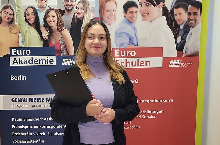 Dozentin Liliia Redko posiert lächelnd für ein Foto. Sie hält ein schwarzes Klemmbrett in der Hand, trägt ein veilchenfarbenen Rollkragenpulli, einen schwarzen Cardigan darüber, eine dünne goldene Halskette und die blonden schulterlangen Haare offen. Im Hintergrund stehen zwei große Roll-Ups der Euro-Schulen und der Euro Akademie.