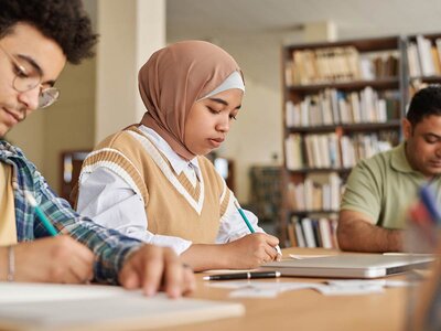 Eine junge Frau mit einem beigefarbenen Kopftuch und einer beigefarbenen Weste sitzt konzentriert an einem Tisch und schreibt auf ein Blatt Papier. Sie hält einen Stift in der Hand und wirkt nachdenklich. Neben ihr sitzen zwei weitere Personen: Ein junger Mann mit Brille und kariertem Hemd sowie ein älterer Mann in einem grünen Polohemd. Im Hintergrund sind Regale mit Büchern zu sehen, was auf eine Lern- oder Arbeitsumgebung, wie eine Bibliothek oder ein Klassenzimmer, hindeutet.