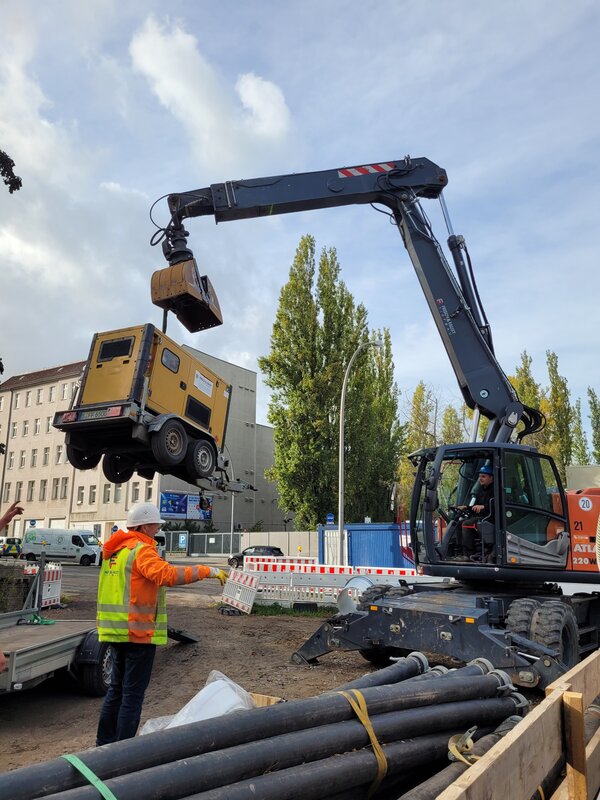 Baustellenszene mit einem großen Bagger, der einen Anhänger anhebt. Ein Mitarbeiter in Warnkleidung weist den Maschinenführer ein, während Absperrungen und Baumaterialien die Baustelle umgeben.