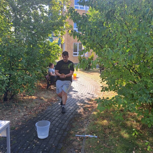 Ein Schüler läuft mit einem nassen Schwamm von einem mit Wasser gefüllten Eimer zu einem anderen mit Wasser gefüllten Eimer. Der Weg, auf dem er läuft, ist von Sträuchern umgeben. Im Hintergrund sitzt eine Schülerin auf einen Parkbank.