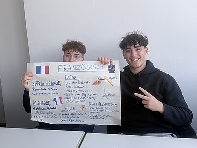 Zwei junge Schüler sitzen im Klassenraum an einem Tisch und posieren für ein Bild. Ein Schüler zeigt gut gelaunt und lächelnd das Victory-Zeichen, der Schüler neben ihm hält dabei ihr Arbeitsblatt zum Thema französische Sprache hoch bis zu seiner Nase, sodass man noch seine Augen, die Brille und die Haare sieht. 