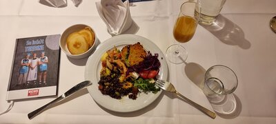 Auf einem weiß gedeckten Tisch steht ein Teller mit einem festlichen Essen, das aus einem gebratenen Stück Fleisch oder Fisch, einer Portion Rotkohl, gemischtem Gemüse und einem Salat besteht. Daneben befindet sich eine kleine Schale mit mehreren Brotscheiben. Auf dem Tisch stehen außerdem ein Glas mit einem hellbraunen Getränk, ein Wasserglas und eine gefaltete weiße Serviette. Links neben dem Teller liegt ein Messer, rechts eine Gabel. Im Hintergrund ist eine aufgestellte Broschüre mit dem Titel 'Sei dabei! TURBINE' und einem Bild von drei Fußballspielerinnen des 1. FFC Turbine Potsdam zu sehen. Das Bild entstand im Rahmen eines Festessens, das vom Standortleiter Islam Shabanah bei den Euro-Schulen Potsdam gesponsert wurde.