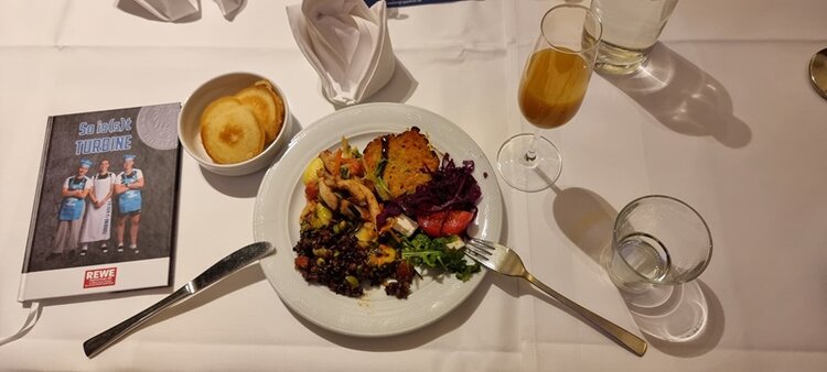 Auf einem weiß gedeckten Tisch steht ein Teller mit einem festlichen Essen, das aus einem gebratenen Stück Fleisch oder Fisch, einer Portion Rotkohl, gemischtem Gemüse und einem Salat besteht. Daneben befindet sich eine kleine Schale mit mehreren Brotscheiben. Auf dem Tisch stehen außerdem ein Glas mit einem hellbraunen Getränk, ein Wasserglas und eine gefaltete weiße Serviette. Links neben dem Teller liegt ein Messer, rechts eine Gabel. Im Hintergrund ist eine aufgestellte Broschüre mit dem Titel 'Sei dabei! TURBINE' und einem Bild von drei Fußballspielerinnen des 1. FFC Turbine Potsdam zu sehen. Das Bild entstand im Rahmen eines Festessens, das vom Standortleiter Islam Shabanah bei den Euro-Schulen Potsdam gesponsert wurde.