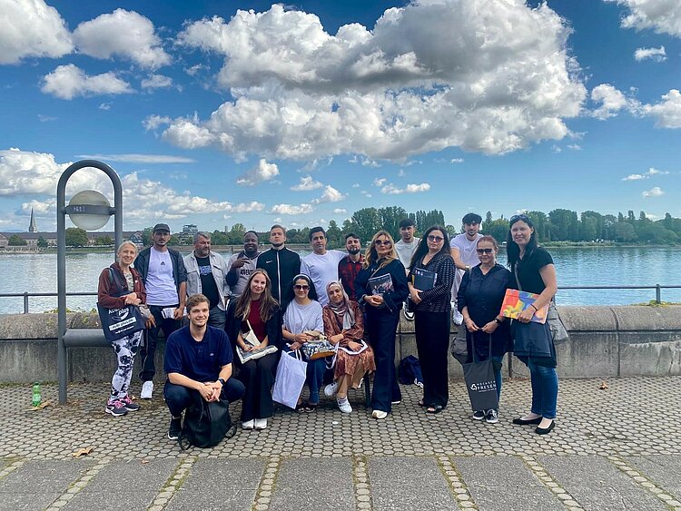 Berufssprachkurs B2 besucht größte Bildungsmesse in der Region Eine Gruppe von etwa 18 Personen steht und sitzt gemeinsam für ein Foto am Rheinufer in Mainz. Im Hintergrund sind der Fluss, Bäume und eine Kirche zu sehen. Die Teilnehmenden lächeln in die Kamera und halten Taschen sowie Unterlagen in den Händen.