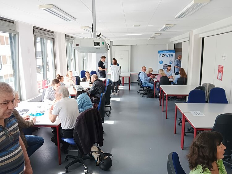 Job-Speed-Dating vom 22.07.2025 ein voller Erfolg! Ein großer Seminarraum voller Menschen, die in Gruppen an mehreren Tischen arbeiten. Im Hintergrund ist ein Roll-Up mit der Aufschrift 'POWER' sichtbar. Tageslicht fällt durch große Fenster in den Raum, der eine offene und aktive Arbeitsatmosphäre zeigt.