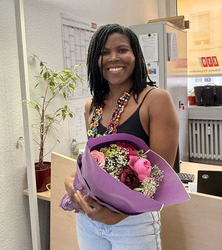 Eine junge Mitarbeitende mit dunkler Haut und schulterlangen, geflochtenen Haaren steht in einem Büro und lächelt in die Kamera. Sie trägt ein schwarzes Oberteil und eine bunte Halskette aus großen Perlen. In den Händen hält sie einen Blumenstrauß mit rosa und violetten Blüten, der in lilafarbenes Papier gewickelt ist. Im Hintergrund sind ein Schreibtisch, ein Computerbildschirm, eine Topfpflanze und eine Wand mit einem Kalender sowie weiteren Unterlagen zu sehen. Durch eine Glastür fällt Tageslicht in den Raum.