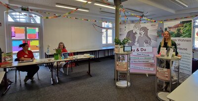 Ein heller Raum mit bunten Papiergirlanden und dekorierten Fenstern. Drei Frauen stehen bzw. sitzen an Info-Tischen, die zur Hausmesse dekoriert sind. Im Hintergrund: ein Roll-up mit der Aufschrift „Du & Du – Zwei Freunde“ der Hera Residenzen.