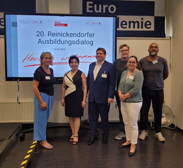 Mitglieder des Reinickendorfer Ausbildungsdialogs posieren für ein Gruppenfoto, im Hintergrund ein großer Bildschirm, auf dem die Teilnehmenden zum 20. RAD willkommen geheißen werden.