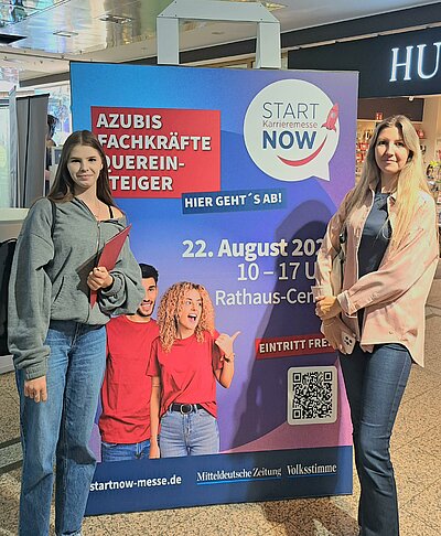 Teilnehmer*innen des Bewerbungscoachings auf der Karrieremesse Start Now in Dessau-Roßlau Zwei junge Frauen stehen vor einem Werbebanner der Karrieremesse START NOW. Auf dem Plakat steht: „Azubis, Fachkräfte, Quereinsteiger – hier geht’s ab! START NOW Karrieremesse, 22. August 2025, 10–17 Uhr, Rathaus-Center, Eintritt frei.“ Unten sind Logos der Mitteldeutschen Zeitung und der Volksstimme zu sehen sowie ein QR-Code.
