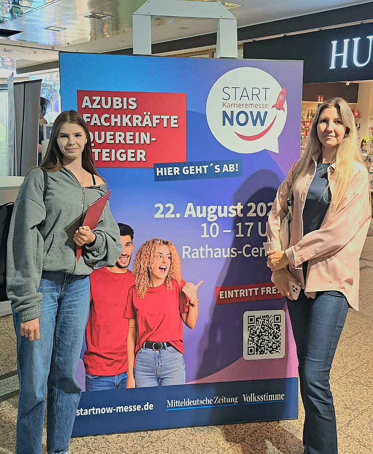 Teilnehmer*innen des Bewerbungscoachings auf der Karrieremesse Start Now in Dessau-Roßlau Zwei junge Frauen stehen vor einem Werbebanner der Karrieremesse START NOW. Auf dem Plakat steht: „Azubis, Fachkräfte, Quereinsteiger – hier geht’s ab! START NOW Karrieremesse, 22. August 2025, 10–17 Uhr, Rathaus-Center, Eintritt frei.“ Unten sind Logos der Mitteldeutschen Zeitung und der Volksstimme zu sehen sowie ein QR-Code.
