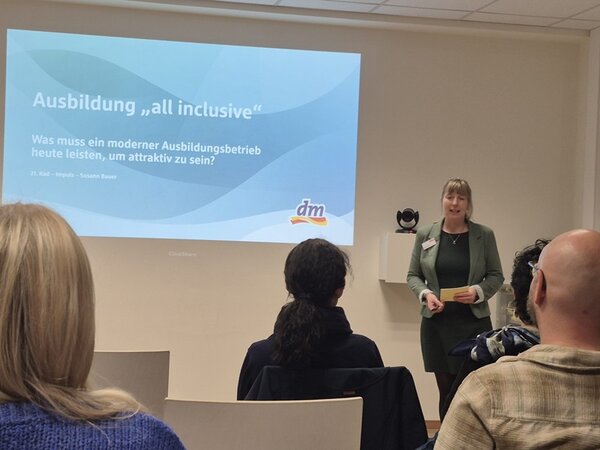 Eine Frau hält einen Vortrag vor einem Publikum und steht rechts neben einer projizierten Präsentationsfolie mit dem Titel ‚Ausbildung all inclusive – Was muss ein moderner Ausbildungsbetrieb heute leisten, um attraktiv zu sein?‘. Zuhörer sitzen im Vordergrund, während die Vortragende in einem grünen Blazer spricht. Die Szene spielt in einem hellen Seminarraum während des 21. Reinickendorfer Ausbildungsdialogs an den Euro-Schulen Berlin.