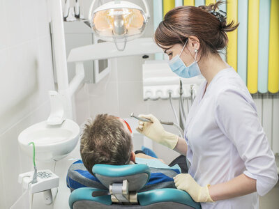Eine Zahnärztin oder zahnmedizinische Fachangestellte in weißer Arbeitskleidung und mit blauer Gesichtsmaske steht neben einem Patienten und führt mit einem zahnärztlichen Instrument eine Behandlung durch. Der Patient sitzt auf einem blauen Behandlungsstuhl und hat den Mund geöffnet. Im Hintergrund sind typische Elemente einer Zahnarztpraxis sichtbar, darunter eine Behandlungslampe, ein Spülbecken und verschiedene zahnärztliche Geräte. Die Atmosphäre wirkt professionell und hygienisch, wie in medizinischen Einrichtungen üblich.