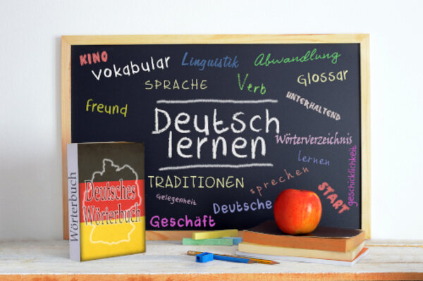 Deutsches Wörterbuch lehnt an einer Tafel, auf der "Deutsch lernen" und weitere deutsche Wörter stehen.