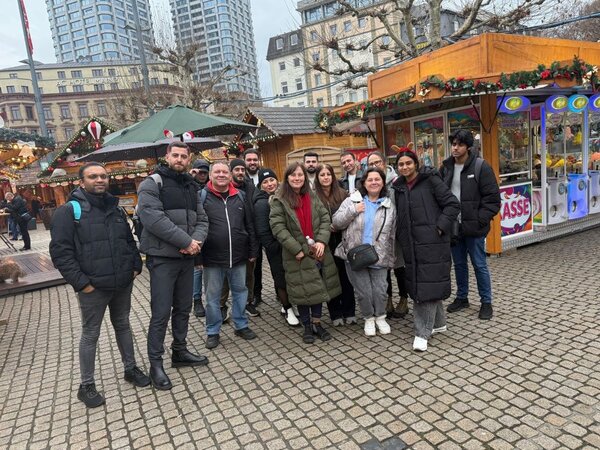 Eine Gruppe von 16 lächelnden Personen steht vor einem festlich dekorierten Stand auf einem Weihnachtsmarkt. Die Personen sind warm angezogen und posieren gemeinsam für ein Foto. Im Hintergrund sind weitere Holzhütten und ein großes Gebäude zu sehen, was auf eine winterliche und gemütliche Atmosphäre hinweist.