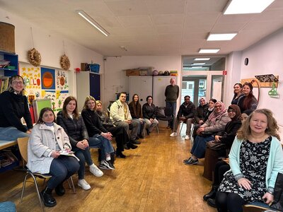 Mehrere erwachsene Kursteilnehmende der Euro-Schulen Wiesbaden sitzen in einem hellen Raum auf Stühlen im Halbkreis. Die Wände sind mit bunten Zeichnungen und Schriftzügen dekoriert. Im Hintergrund stehen Regale, Schränke und eine Pinnwand. Der Boden besteht aus Holz. Einige Personen tragen Jacken, andere Pullover oder Schals. Die Stimmung wirkt ruhig und konzentriert.