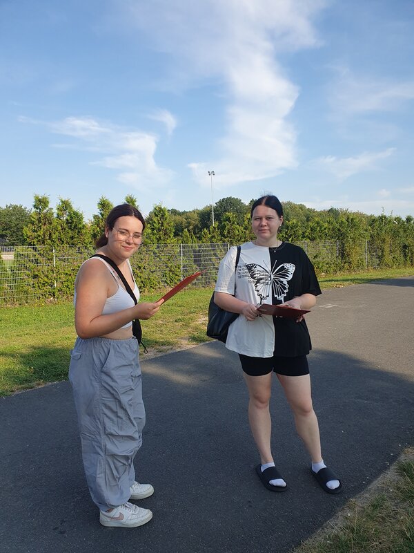 Zwei Schülerinnen in sommerlicher Kleidung stehen im Schatten auf einem asphaltierten Fahrradweg, dahinter befindet sich ein Sportplatz. Beide Mädchen halten eine Mappe in der Hand und haben eine Tasche umhängen.
