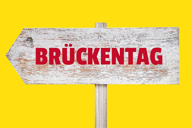 Wichtiger Hinweis in eigener Sache! Ein weiß gestrichenes Holzschild in Pfeilform mit roter Aufschrift „Brückentag“, vor einem leuchtend gelben Hintergrund.