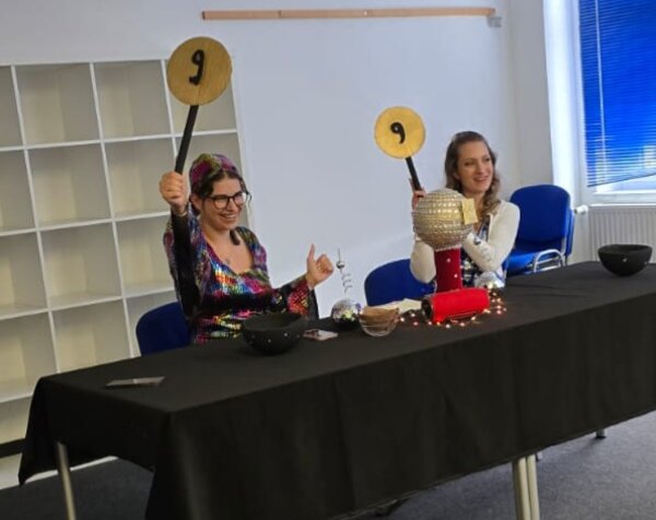 Zwei Personen sitzen an einem Jury-Tisch und halten Bewertungstafeln mit der Zahl 9 hoch.