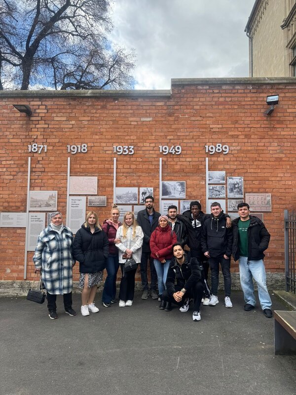 Vor einer roten Backsteinmauer mit historischen Jahreszahlen und erklärenden Texttafeln steht die Gruppe der Teilnehmenden des Integrationskurses. Die Jahreszahlen 1871, 1918, 1933, 1949 und 1989 sind in großen Ziffern oberhalb der Tafeln angebracht. Die Personen sind unterschiedlich gekleidet, einige stehen, andere hocken, und sie blicken in Richtung Kamera. Im Hintergrund sind kahle Äste eines Baumes und ein Teil eines Gebäudes zu sehen. Der Himmel ist bewölkt.