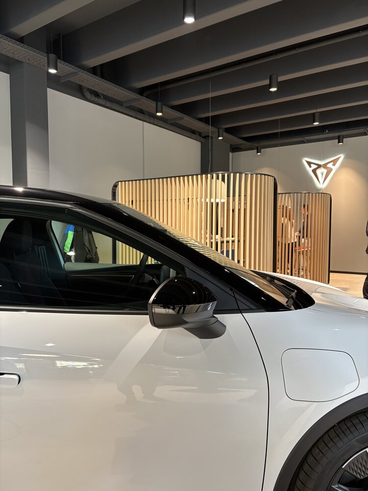 Detailaufnahme eines weißen CUPRA-Fahrzeugs in einem modernen Autohaus. Im Hintergrund sind holzverkleidete Beratungsbereiche und das beleuchtete CUPRA-Logo an der Wand zu sehen.