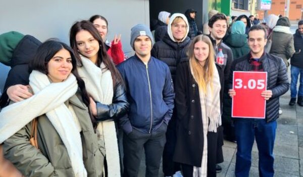 Eine Gruppe von neun Personen posiert draußen in einer Menschenmenge. Zwei Frauen im Vordergrund tragen weiße Schals. Ein Mann in der Gruppe hält ein Schild mit der roten Aufschrift „3.05“. Alle Personen tragen Winterkleidung, darunter Jacken und Mützen. Weitere Menschen sind im Hintergrund zu sehen, ebenfalls warm angezogen.