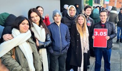 Eine Gruppe von neun Personen posiert draußen in einer Menschenmenge. Zwei Frauen im Vordergrund tragen weiße Schals. Ein Mann in der Gruppe hält ein Schild mit der roten Aufschrift „3.05“. Alle Personen tragen Winterkleidung, darunter Jacken und Mützen. Weitere Menschen sind im Hintergrund zu sehen, ebenfalls warm angezogen.