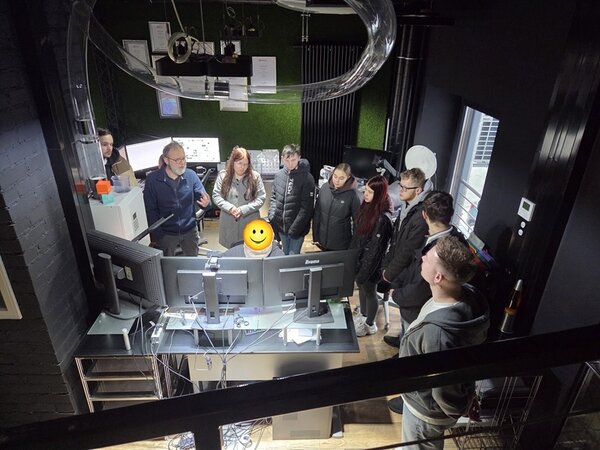 Mehrere Auszubildende der Euro-Schulen Hoyerswerda stehen in einem modernen Büroraum einer Werbeagentur. Die Gruppe ist um einen Arbeitsplatz mit mehreren Monitoren und technischen Geräten versammelt. Im Hintergrund sind grüne Wandpaneele und technische Installationen an der Decke sichtbar. Die Auszubildenden tragen überwiegend legere Kleidung wie Jacken und Jeans. Die Szene vermittelt einen Einblick in den Arbeitsalltag einer Werbeagentur und dokumentiert den Besuch der angehenden Verkäuferinnen und Verkäufer im Rahmen ihrer Ausbildung.