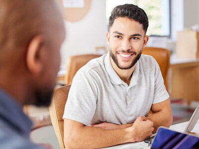 Ein junger Mann mit kurzem, dunklem Haar und Bart sitzt lächelnd an einem Tisch in einem Büro oder Besprechungsraum. Er trägt ein hellgraues Poloshirt und wirkt entspannt und freundlich. Vor ihm auf dem Tisch liegt ein Tablet oder ein Notizblock, und er hat die Arme verschränkt. Im Hintergrund ist eine andere Person zu sehen, deren Gesicht nicht erkennbar ist. Der Raum wirkt hell und einladend, mit natürlichem Licht, das durch ein Fenster fällt. Die Szene vermittelt den Eindruck eines positiven Gesprächs oder einer Besprechung.