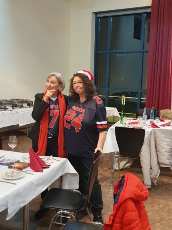 Zwei Frauen stehen nebeneinander an einem gedeckten Tisch und lächeln in die Kamera. Beide tragen Football-Trikots in dunklen Farben, eine hat zusätzlich eine rote Weihnachtsmütze auf, im Hintergrund sind weitere Tische und ein Buffet aufgebaut.