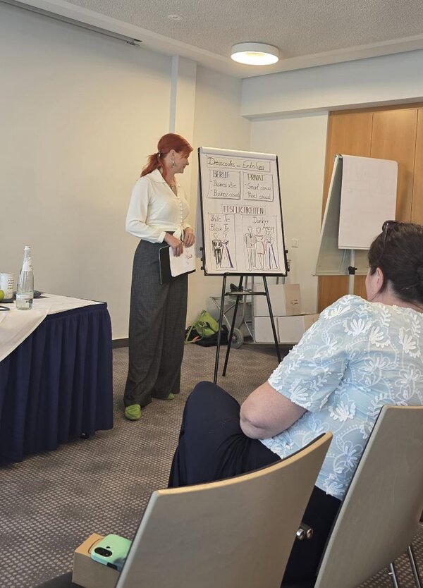 Eine Frau mit roten Haaren und einem weißen Oberteil steht vor einem Flipchart, auf dem Skizzen von Kleidungsstücken und Notizen zu sehen sind. Sie scheint eine Präsentation zu halten. Im Vordergrund sitzt eine Person mit dem Rücken zur Kamera.