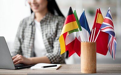 Berufssprachkurs Frühpädagogik B2 – jetzt bei den Euro-Schulen Oldenburg Eine Frau sitzt an einem Notebook und tippt dort ein. Rechts davor steht ein Gefäß mit verschiedenen Landesflaggen, die darin stecken.