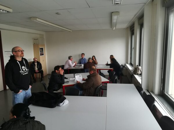 Menschen beraten sich in kleinen Gruppen an Tischen; im Vordergrund ein leerer Tisch, rechts Fenster mit Blick nach draußen.