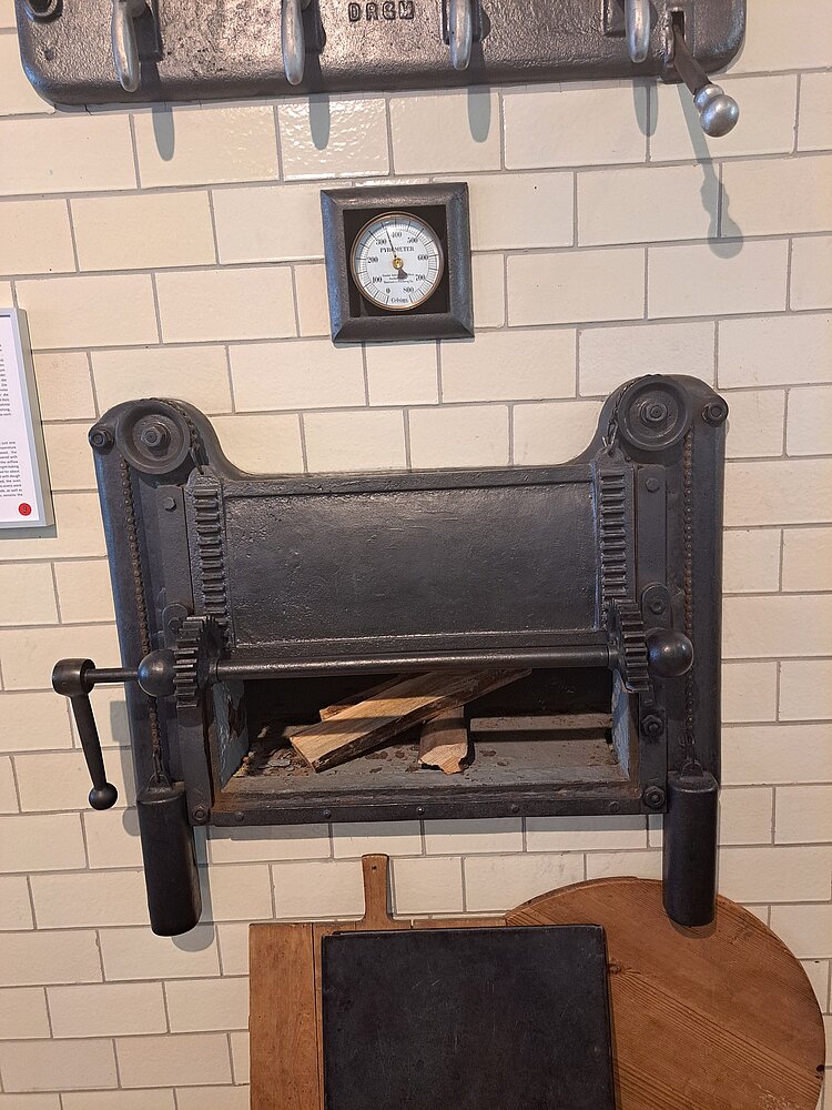 Nahaufnahme eines historischen Ofens mit Thermometer und Holzscheiten im Brennraum. Der Ofen ist in eine weiß geflieste Wand eingebaut und zeigt schwere Metallteile mit Zahnrädern und Griffen.