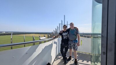 Auf einem Aussichtsturm stehen zwei Jungs, die für das Foto die Arme umeinander gelegt haben. Im Hintergrund sind Windräder erkennbar, der Himmel ist wolkenlos.