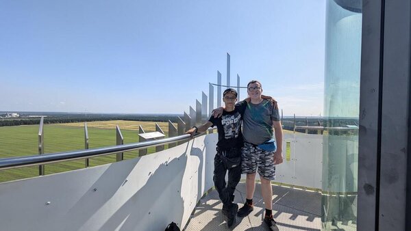 Auf einem Aussichtsturm stehen zwei Jungs, die für das Foto die Arme umeinander gelegt haben. Im Hintergrund sind Windräder erkennbar, der Himmel ist wolkenlos.